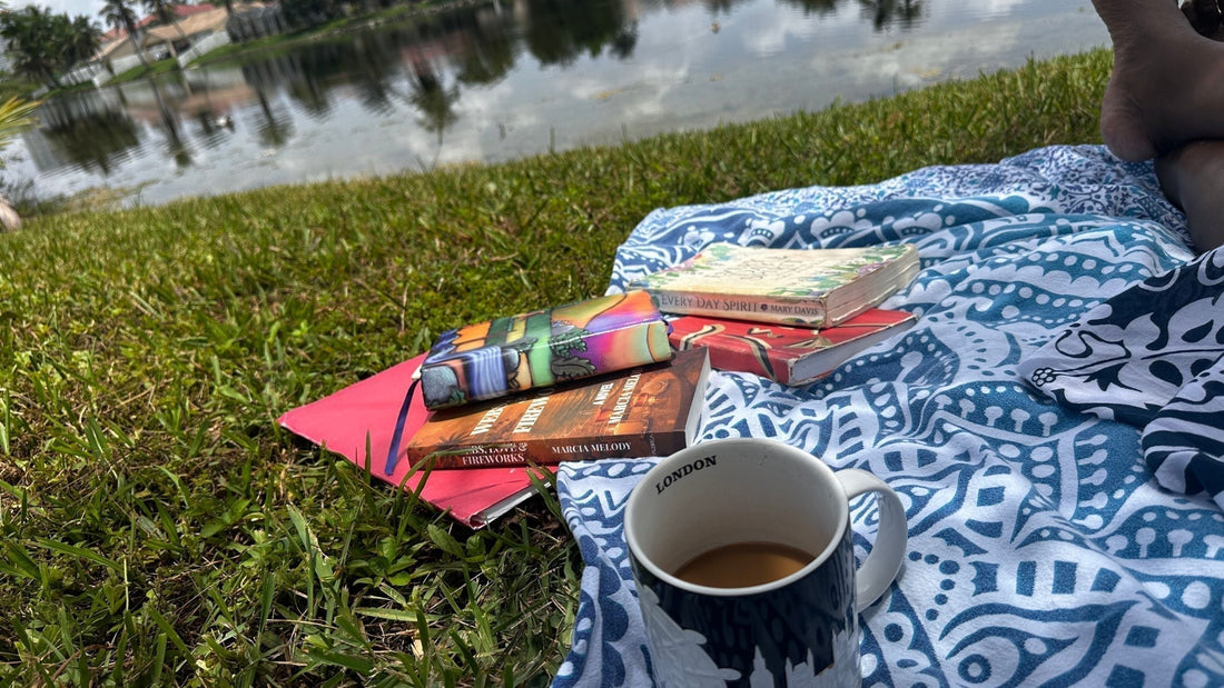 First-person view from a blue patterned picnic blanket by a calm Florida lake. A London coffee mug sits next to a stack of books featuring the novel Webs, Love & Fireworks by author Marcia Melody.