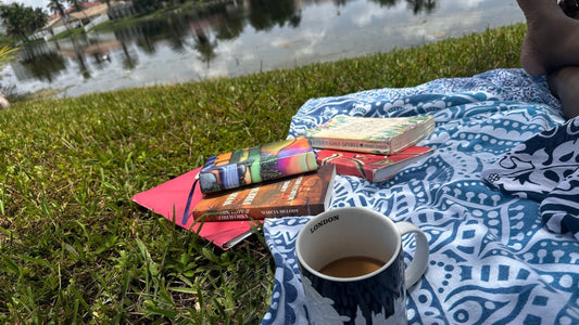 First-person view from a blue patterned picnic blanket by a calm Florida lake. A London coffee mug sits next to a stack of books featuring the novel Webs, Love & Fireworks by author Marcia Melody.