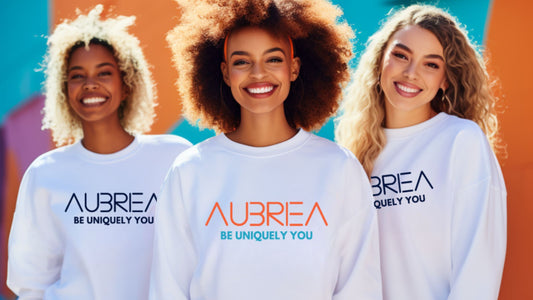 Joyful group of diverse women laughing and celebrating in unity, dressed in Aubriea's signature white sweatshirts with the uplifting slogan 'Be Uniquely You' emblazoned in orange. 