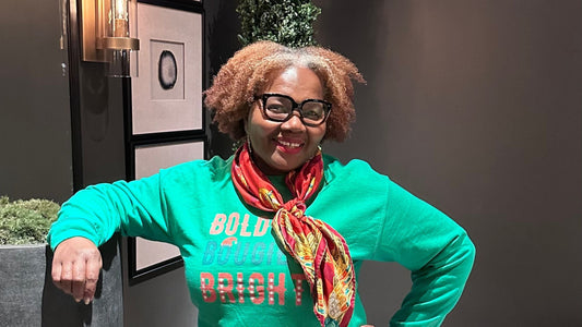 Marcia Allen, the founder of NASTÉ Apparel radiates joy and confidence in her signature 'Bold, Bougie, and Bright' sweatshirt. The vivacious green hue of the sweatshirt pops against the elegant, dark-toned backdrop of a stylishly designed room.