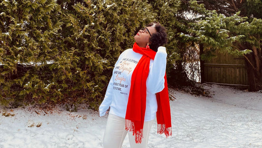A woman in a NASTÉ Apparel white sweatshirt with "she is clothed in strength and dignity" stands against a wintry backdrop, her pose reflective and uplifted, with a red scarf and boots, embodying the brand's message of empowerment and positivity.